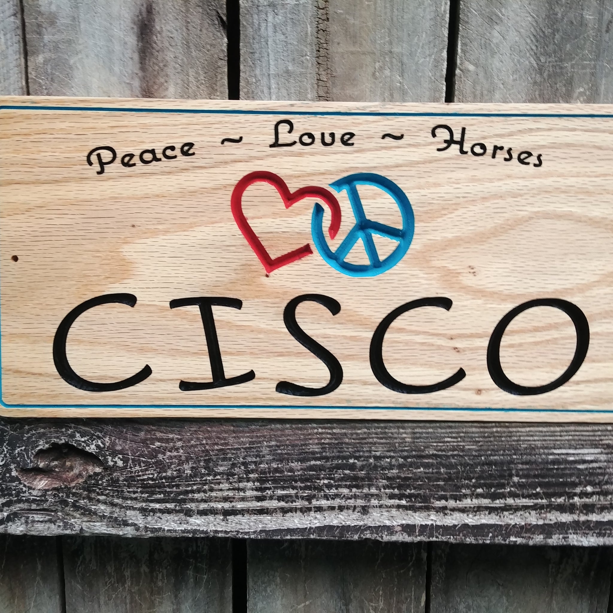 This is a personalized solid Red Oak Horse stall sign. Custom made signage with Horse name in a whimsical font, an interlocking red heart and blue peace sign and the words peace-love-horses at the top. All text is v-carved into the solid wood and painted.