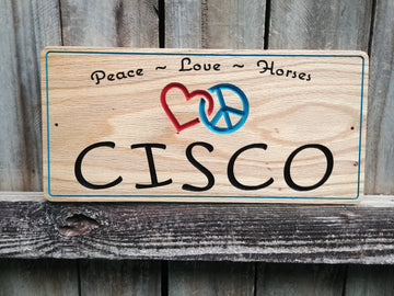 This is a personalized solid Red Oak Horse stall sign. Custom made signage with Horse name in a whimsical font, an interlocking red heart and blue peace sign and the words peace-love-horses at the top. All text is v-carved into the solid wood and painted.